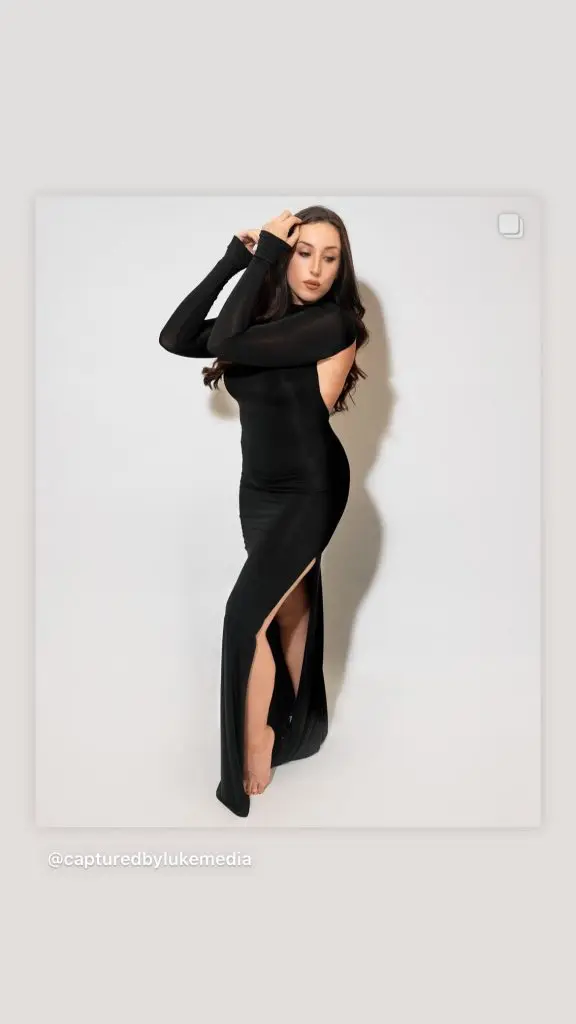Content creator in a long black dress on a white studio backdrop, shown on a phone screen