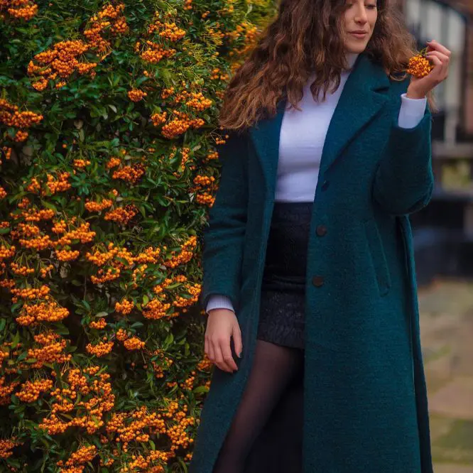 Singer in a teal coat standing beside a wall of orange flowers