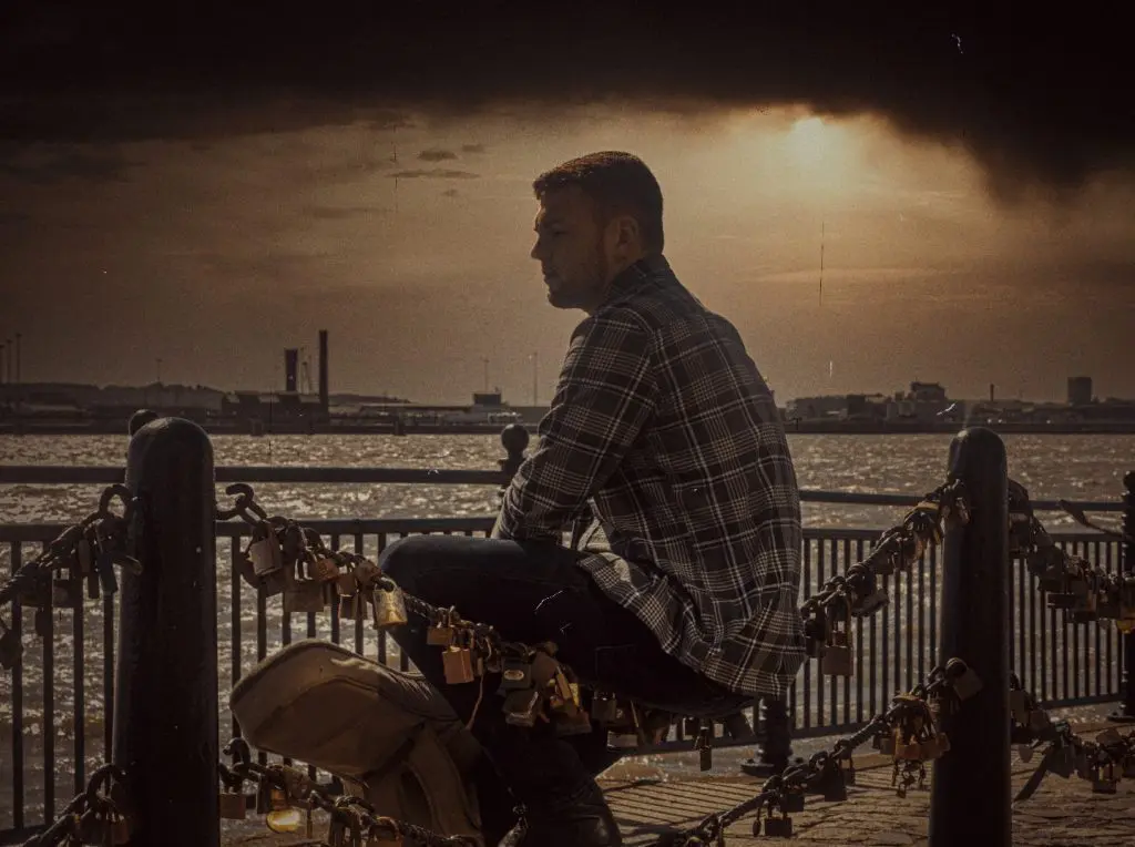 Music artist seated on waterfront railings at golden hour under a dramatic sky