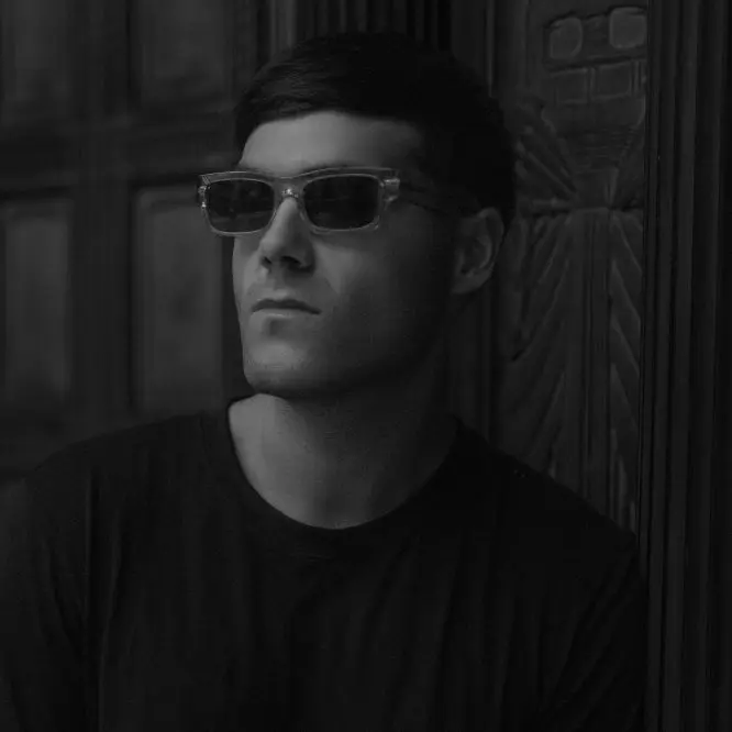 DJ in sunglasses standing in a shadowed doorway lit from one side