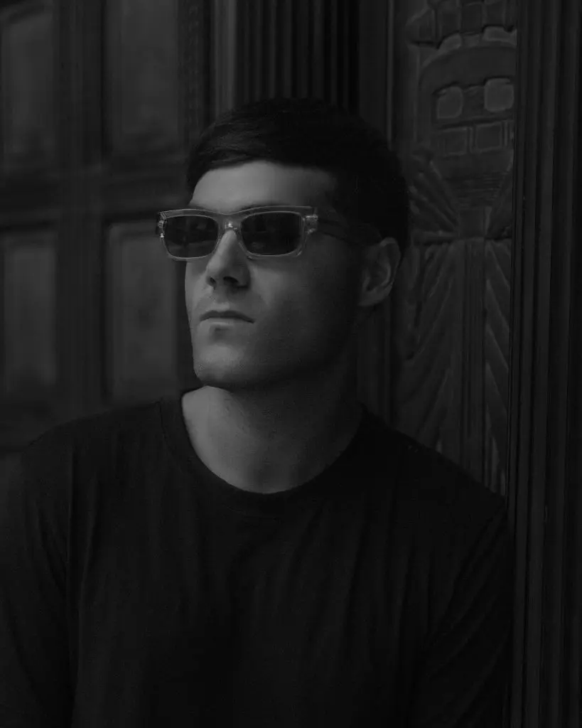 DJ in sunglasses standing in a shadowed doorway lit from one side