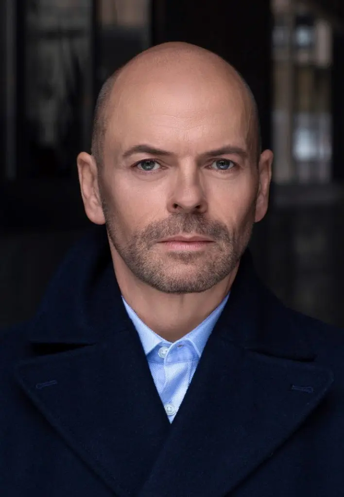 Professional headshot of a man in a navy coat and light blue shirt