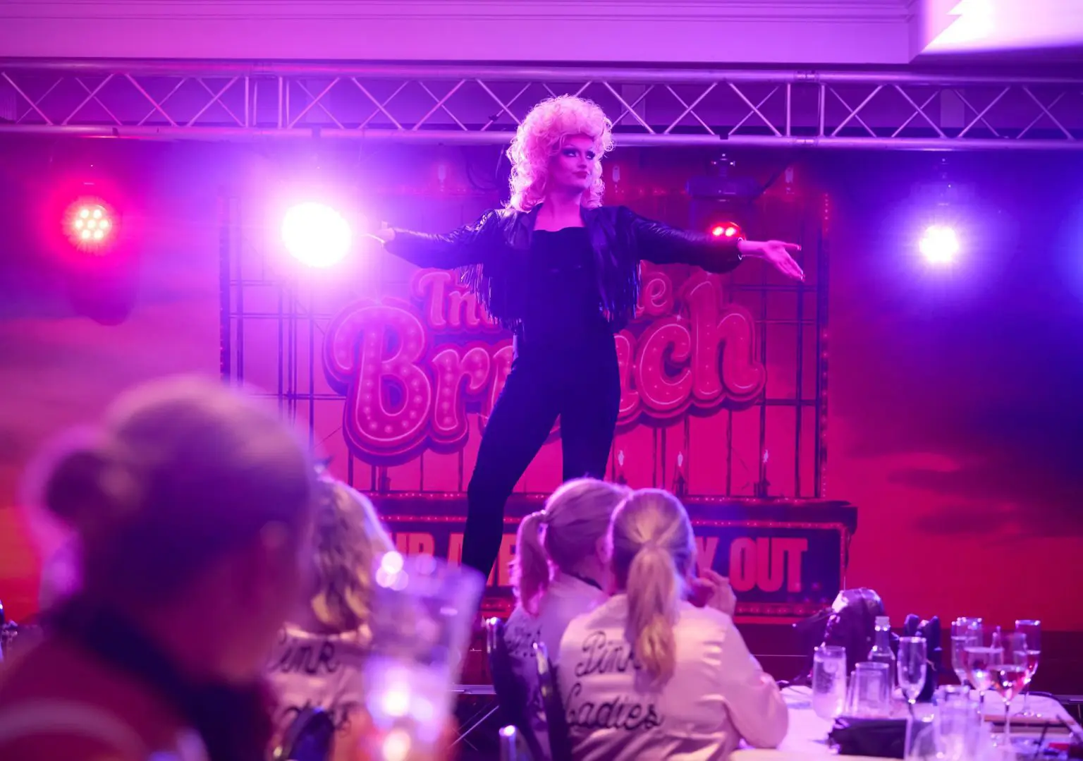 Drag artist performing on stage under pink and purple lights at The Incredible Brunch, a Grease themed bottomless brunch in Liverpool, with guests in Pink Ladies jackets watching