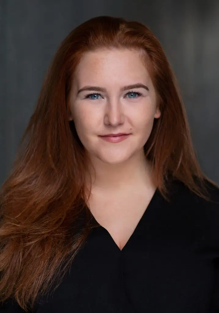 Headshot of a red-haired actress in a black shirt against a dark background
