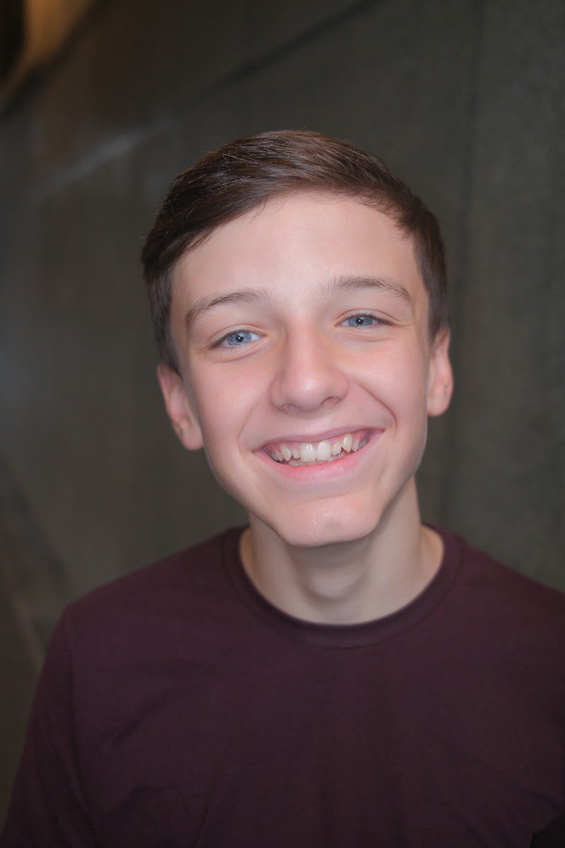 Young actor in a burgundy t-shirt smiling warmly to camera, studio headshot