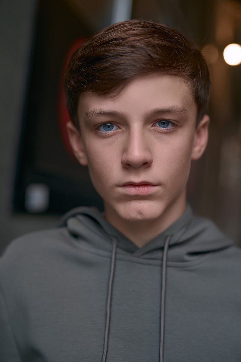 Young actor in a grey hoodie on location in Liverpool, steady gaze with warm lights behind