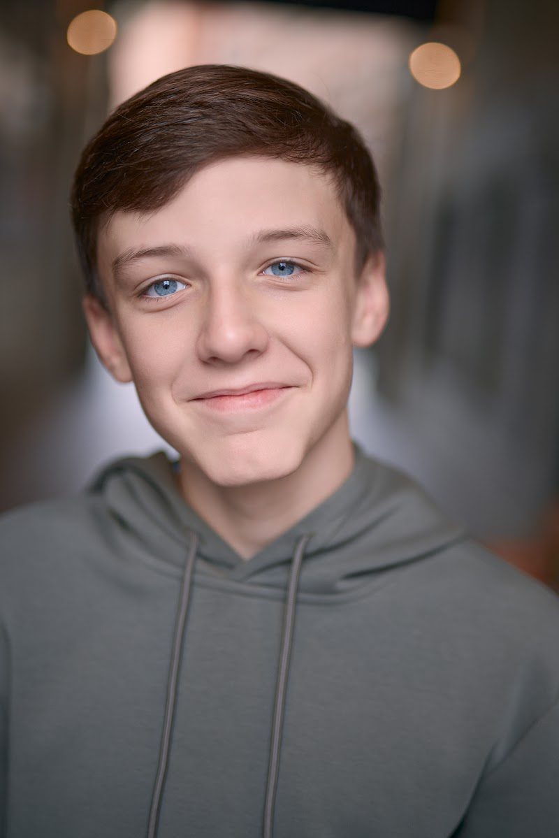 Young actor in a grey hoodie, easy smile with warm city lights behind