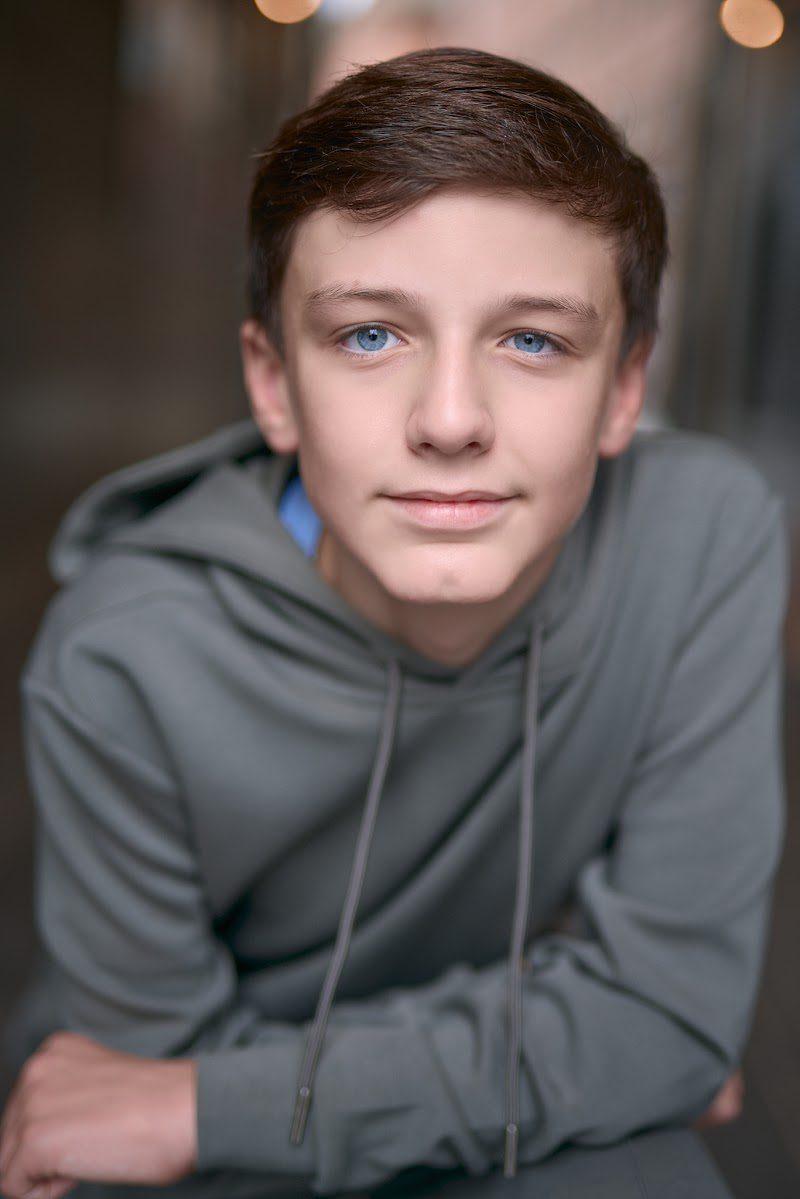 Young actor in a grey hoodie crouched forward with arms on knees, soft smile, on location