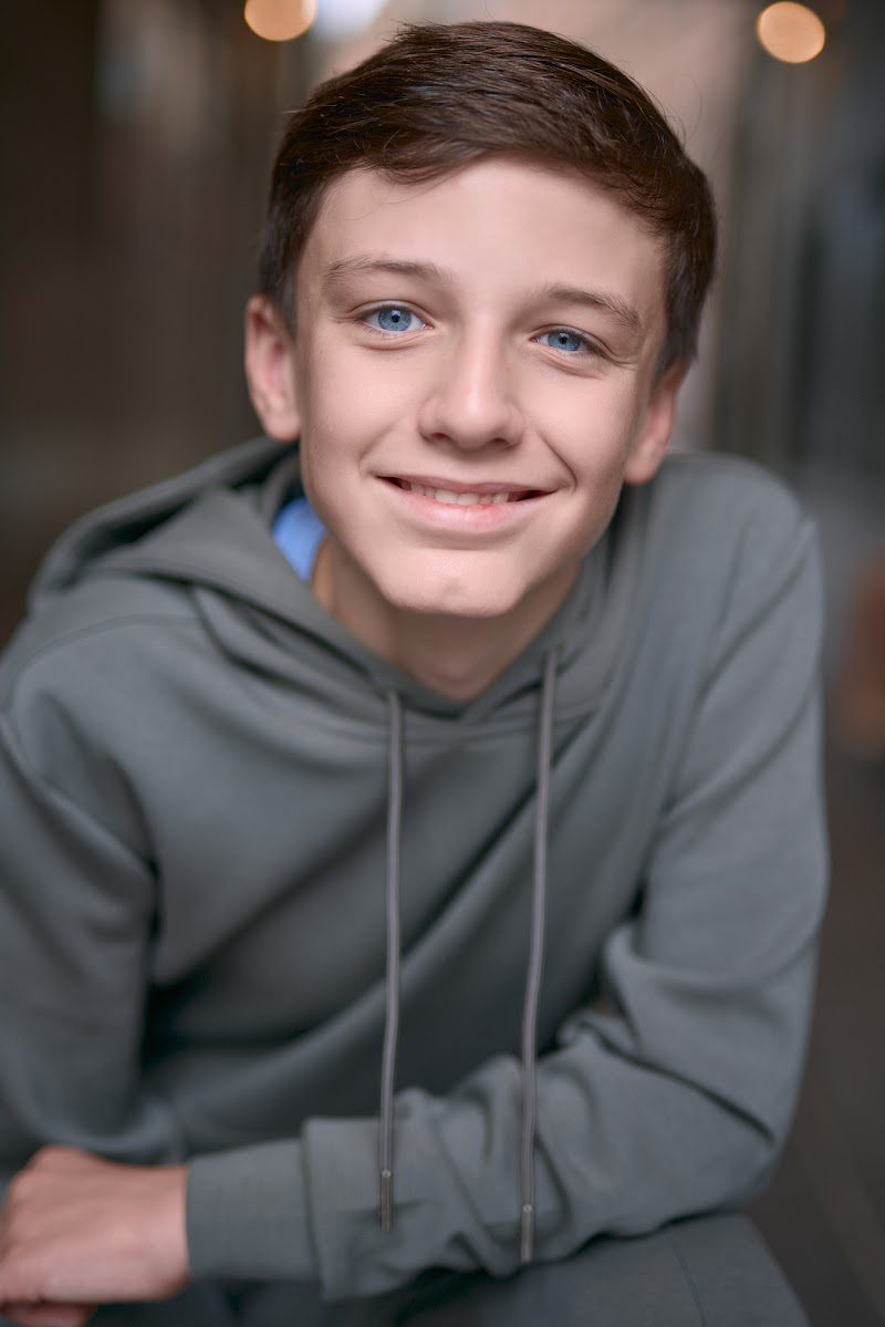 Young actor in a grey hoodie crouched forward with arms on knees, bright smile, on location
