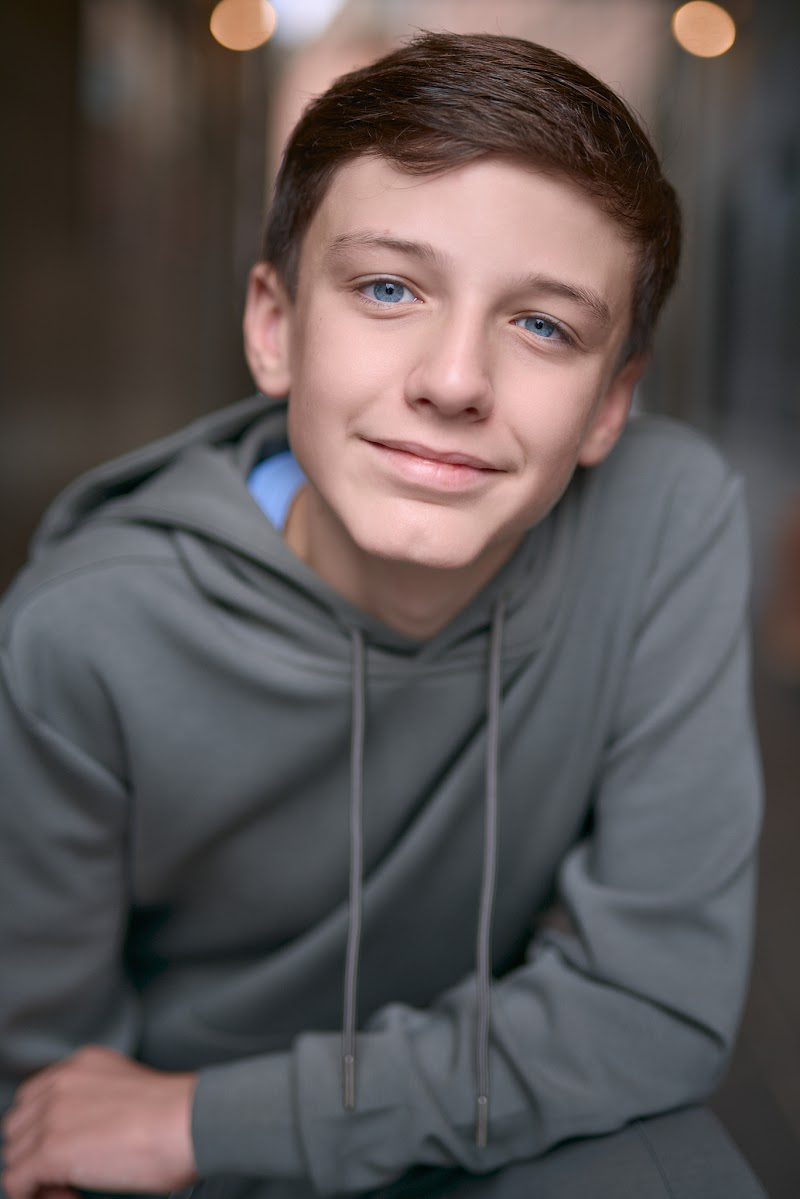 Young actor in a grey hoodie crouched forward with arms on knees, relaxed grin, on location