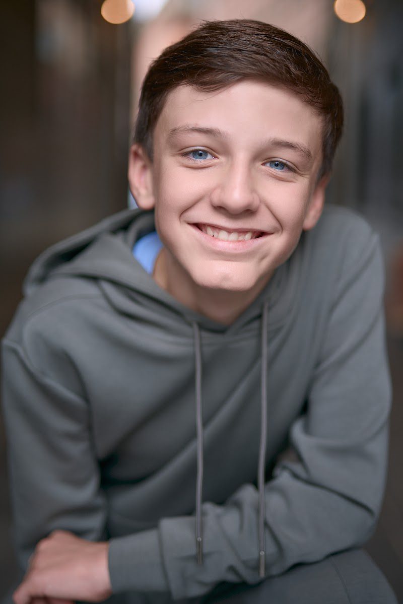 Young actor in a grey hoodie crouched forward with arms on knees, warm smile, eyes to camera, on location