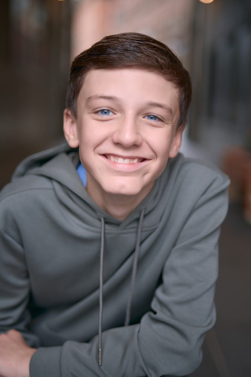 Young actor in a grey hoodie crouched forward with arms on knees, big natural smile, on location