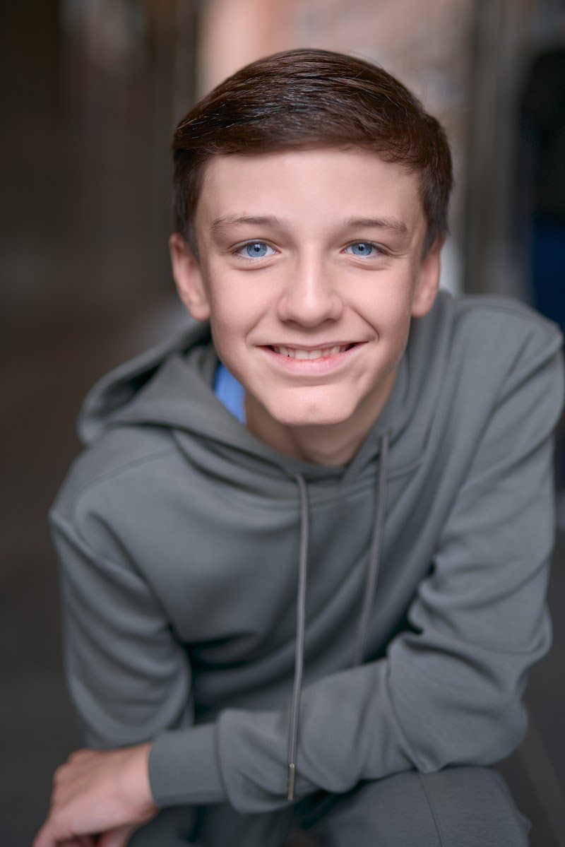 Young actor in a grey hoodie crouched forward with arms on knees, friendly smile, head tilted, on location