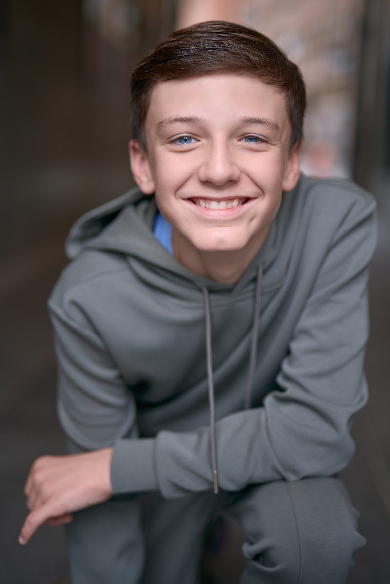 Young actor in a grey hoodie crouched forward with arms on knees, cheeky grin, on location