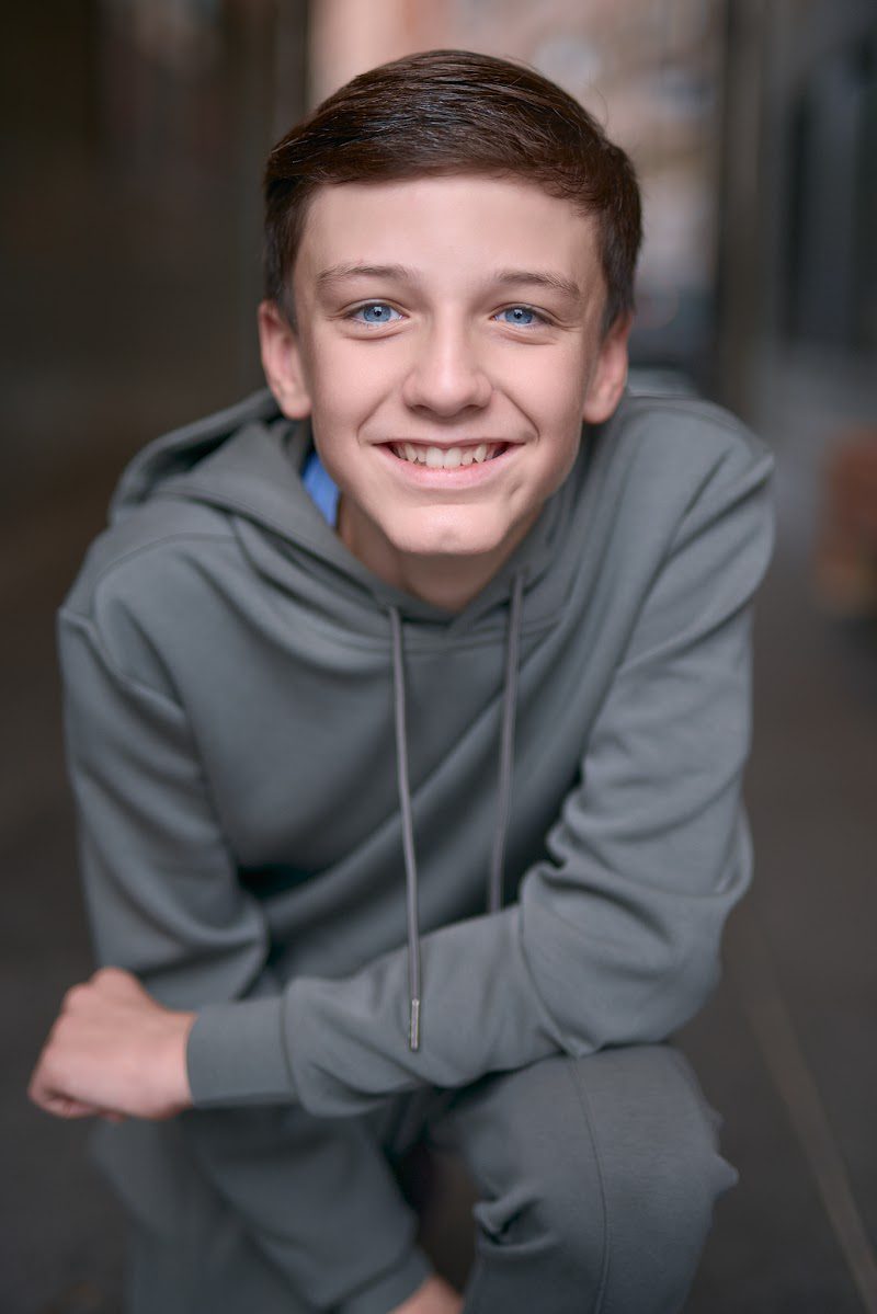 Young actor in a grey hoodie crouched forward with arms on knees, easy smile, on location